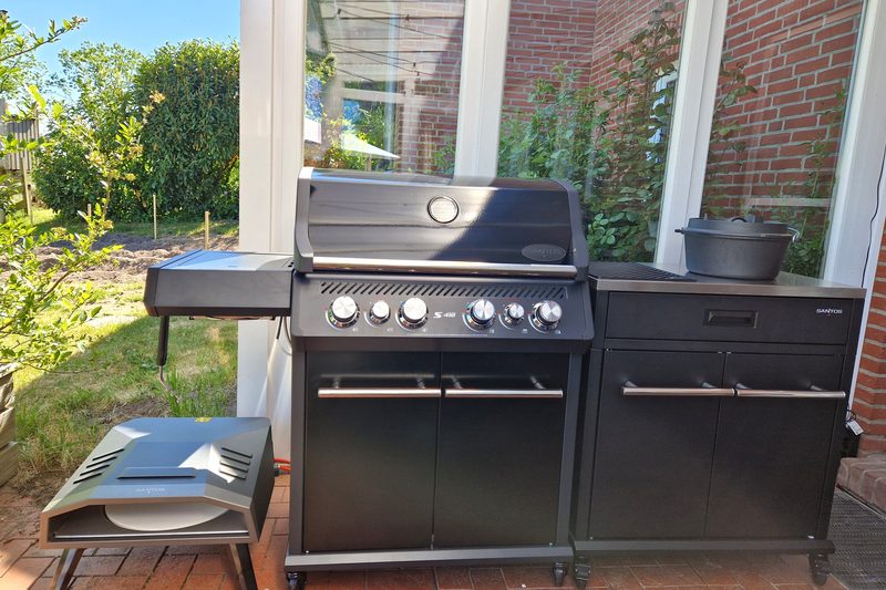 Schwarze Grillstation mit geschlossenem Deckel, links der Seitenkocher, rechts ein Arbeitsmodul mit Gusseisentopf auf Terrassenklinker; vorn links steht der Pizzaofen, der einzeln vermietet wird und nicht zur Grillmiete gehört