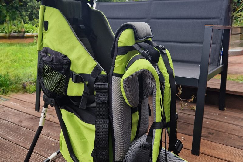 Grüne Rückentrage von der Seite, mit Netztasche, Tragegurten und gepolstertem Hüftgurt, aufgestellt auf einer Holzterrasse