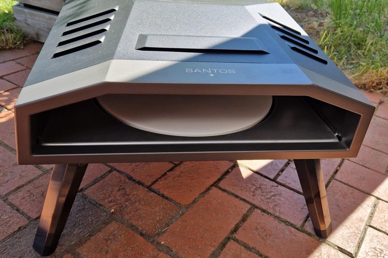 Kompakter Aufsatzofen von vorn, hinter der breiten Öffnung der runde Pizzastein, auf vier schräg gestellten Metallbeinen, auf einer Klinkerterrasse