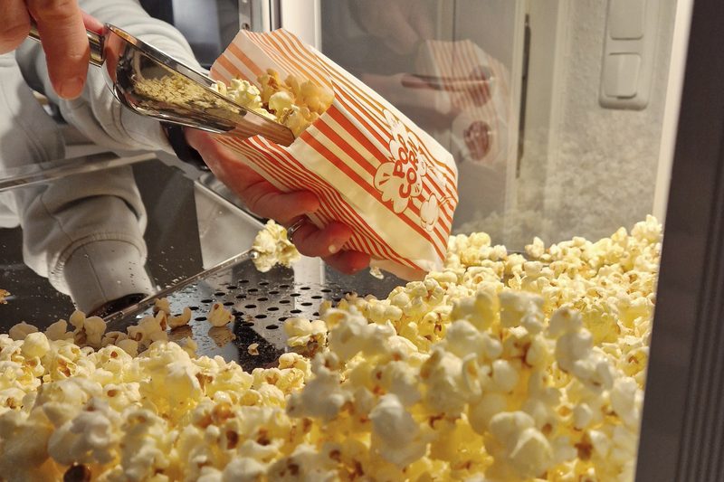 Hand füllt frisches Popcorn mit einer Metallschaufel in eine rot-weiß gestreifte Tüte